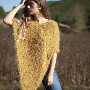May include: A woman wearing a fuzzy, mustard yellow poncho with a long, flowing hairstyle. She is standing in a field of dry grass.