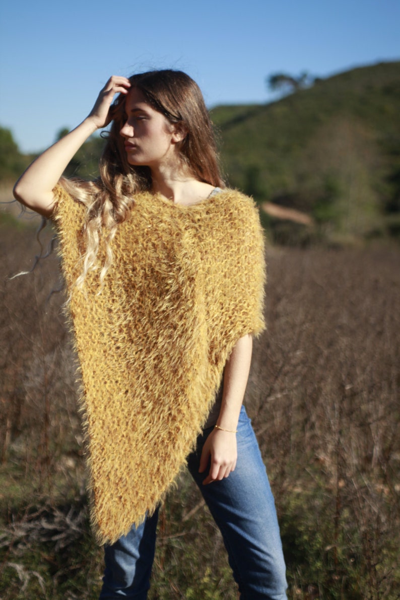 May include: A woman wearing a fuzzy, mustard yellow poncho with a long, flowing hairstyle. She is standing in a field of dry grass.