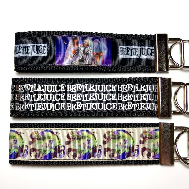 May include: Three black and white keychains with different designs. The top keychain features a picture of the characters from the movie Beetlejuice. The middle keychain has the words "Beetlejuice" repeated multiple times. The bottom keychain has a repeating pattern of a green and purple ghost.