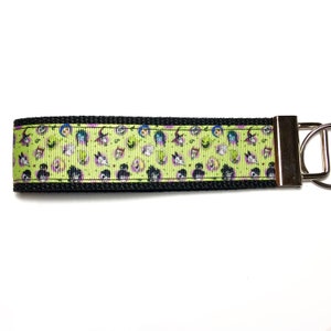 May include: A black and green key chain with a repeating pattern of cartoon faces wearing witch hats. The key chain has a silver metal ring.