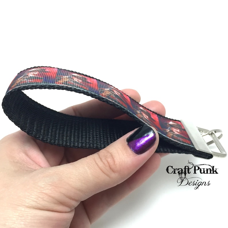 May include: A black and red patterned key chain with a silver metal clasp. The key chain is made of a black woven fabric with a red, white, and black patterned ribbon.