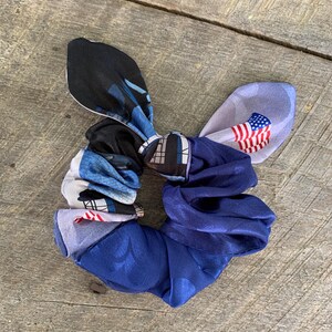 May include: A multi-patterned scrunchie with a bow. The scrunchie features a mix of blue, white, and black fabrics, including a section with an American flag design. The scrunchie is made of a soft, silky material.
