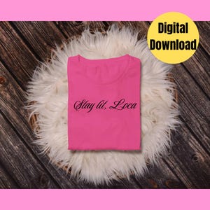 May include: A folded, hot pink t-shirt with the text "Stay lit, Loca" in black script. The shirt is displayed on a white, fluffy surface against a dark wood background. A yellow circle with "Digital Download" is in the upper right corner.