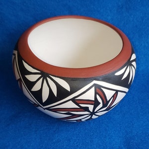 May include: A handcrafted ceramic bowl with a wide, open design. The bowl features a white interior, a dark red rim, and a black band with white and red geometric floral patterns. The bowl sits on a blue surface.