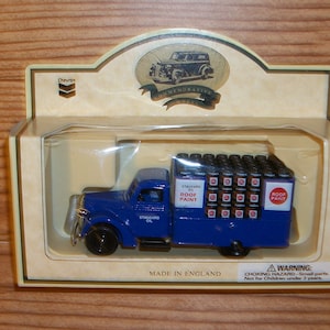 May include: A blue toy truck with a load of roof paint cans, packaged in a commemorative box. The truck has the words "Standard Oil" and "Roof Paint" printed on it. The box is cream-colored with the Chevron logo.