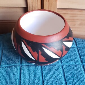 May include: A round, hand-painted ceramic bowl with a wide opening. The bowl has a red-brown exterior with a black and white geometric pattern. The interior is white, and the bowl sits on a blue towel.