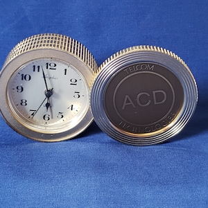 May include: A silver-toned, round travel clock with a light-colored face and black numerals. The clock face reads "Angelus" and "Quartz." The other side of the clock has the text "TELCOM ACD TECHNOLOGIES" in a circular design.