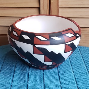 May include: A ceramic bowl with a white interior and a geometric pattern in black, red, and white. The bowl has a rounded shape with a slightly flared rim. The design features angular shapes and lines, creating a Southwestern or Native American aesthetic.