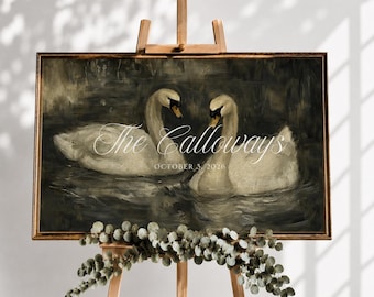 May include: A rectangular framed print featuring an oil painting of two white swans swimming on dark water. The text reads The Calloways and OCTOBER 3, 2026. The print is displayed on a wooden easel decorated with a garland of green eucalyptus leaves.