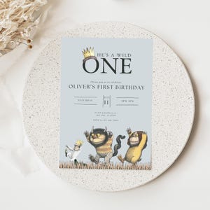 Pode incluir: Um convite de aniversário azul claro com o texto "HE'S A WILD ONE" e "OLIVER'S FIRST BIRTHDAY". O convite apresenta ilustrações de três monstros de desenhos animados e a data de 11 de julho. O convite está em uma superfície branca texturizada.