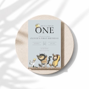 May include: A rectangular birthday invitation card on a light blue background featuring a gold crown icon above the text HE'S A WILD ONE. Below, it reads OLIVER'S FIRST BIRTHDAY, JULY 11, and includes three illustrated monsters standing in grass.