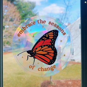 May include: A round sticker with a holographic background featuring a monarch butterfly and the text "embrace the seasons of change".