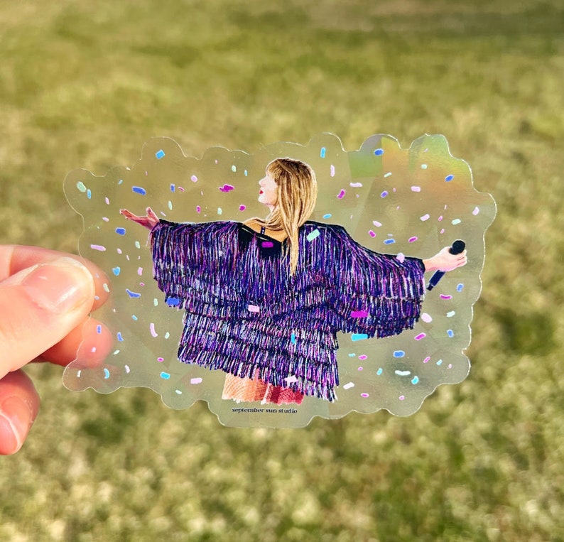 May include: A clear sticker with a holographic finish featuring a woman in a purple sequined jacket with confetti falling around her. The text "september sun studio" is at the bottom of the sticker.