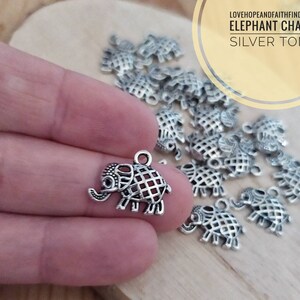 Può includere: Un primo piano di un ciondolo a forma di elefante in tono argento. Il ciondolo è dettagliato con un motivo a griglia e ha un anello per attaccarlo a una collana o un braccialetto. Il ciondolo è circondato da altri ciondoli simili.