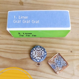 Könnte beinhalten: Ein grüner und blauer Polierblock für Polymer Clay. Der Block hat zwei Seiten, eine mit der Aufschrift "Limer Grat Grat Grat" und die andere mit der Aufschrift "Lisser Zip Zip Zip". Der Block liegt auf einer Holzoberfläche mit zwei kleinen Keramikfliesen.