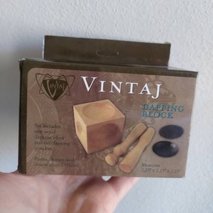 Könnte beinhalten: Ein VINTAJ-Prägeblock-Set, einschließlich eines Holzblocks, zwei Punzen und Formen. Die Box misst 5,7 cm x 5,7 cm x 5,7 cm. Das Set dient zum Formen von Metallrohlingen in Formen, Kuppeln und anderen Formen.