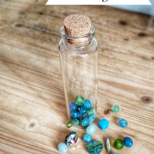 Może przedstawiać: Mała szklana butelka z korkowym korkiem, wypełniona niebieskimi i zielonymi szklanymi koralikami. Butelka stoi na drewnianej powierzchni, a wokół niej rozrzucone są kolejne koraliki.