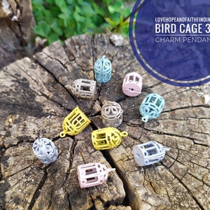 Può includere: Una collezione di ciondoli a forma di gabbia per uccelli 3D colorati, in giallo, rosa, grigio e blu. I ciondoli sono sparsi su una superficie di legno invecchiato. Il testo sull'immagine recita: "LOVEHOPEANDFAITHFINDINGS BIRD CAGE 3D CHARM PENDANT."