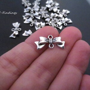 May include: A close-up of a hand holding a silver bow charm. The charm has a loop on each side for attaching to jewelry or other crafts.