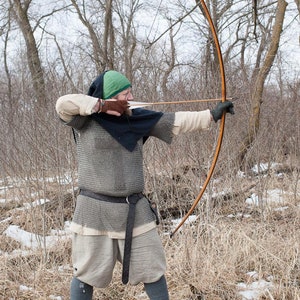 May include: A person wearing a green hooded tunic and chainmail is holding a longbow and arrow, aiming at a target. The person is standing in a snowy forest setting.