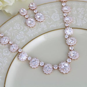 May include: A rose gold necklace and earrings set with clear cubic zirconia stones. The necklace features a delicate chain with large, round stones and smaller stones in between. The earrings are simple studs with round stones.