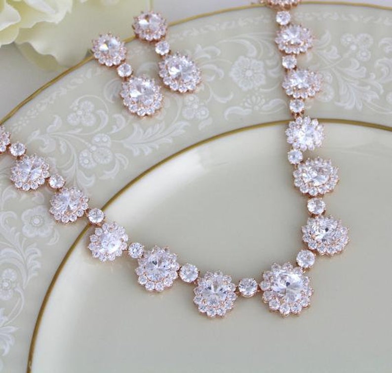 May include: A rose gold necklace and earrings set with clear cubic zirconia stones. The necklace features a delicate chain with large, round stones and smaller stones in between. The earrings are simple studs with round stones.