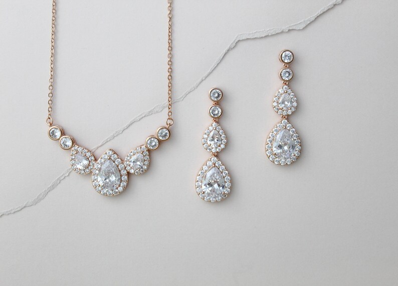 Pu&ograve; includere: Un set di collana e orecchini in oro rosa con pietre di zircone cubico a forma di pera. La collana presenta un gruppo di tre pietre, mentre gli orecchini ne hanno tre ciascuna, a cascata.