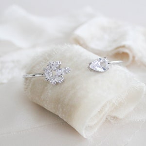 K&ouml;nnte beinhalten: Zwei silberne Ringe mit klaren Zirkonia-Steinen. Ein Ring hat eine Ansammlung von Steinen in einem Blumenmuster, w&auml;hrend der andere Ring einen einzelnen birnenf&ouml;rmigen Stein hat.