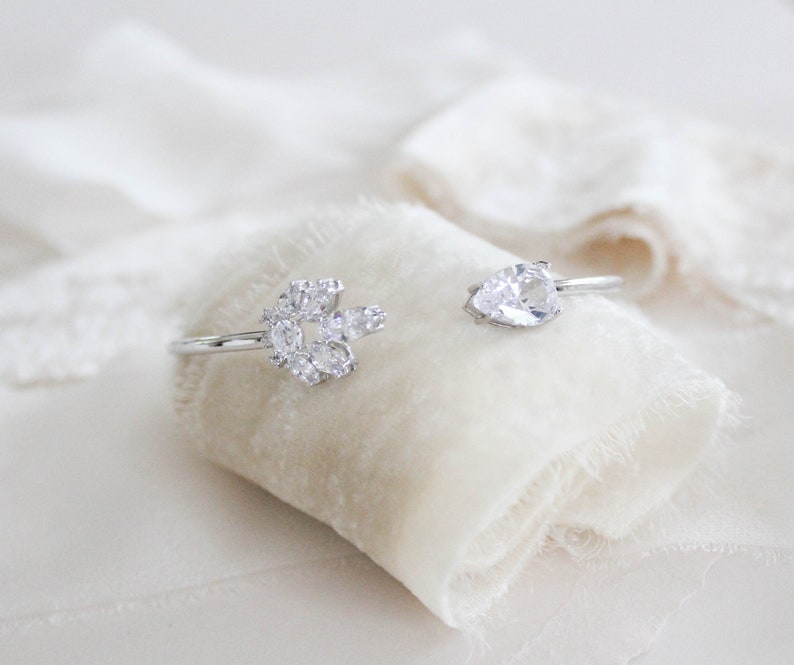K&ouml;nnte beinhalten: Zwei silberne Ringe mit klaren Zirkonia-Steinen. Ein Ring hat eine Ansammlung von Steinen in einem Blumenmuster, w&auml;hrend der andere Ring einen einzelnen birnenf&ouml;rmigen Stein hat.