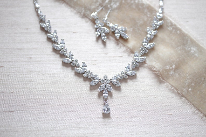 May include: A silver necklace and earrings set with a teardrop pendant and a delicate leaf design. The jewellery is made with clear cubic zirconia stones.