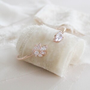 K&ouml;nnte beinhalten: Ein ros&eacute;goldenes Armband mit einem Cluster aus klaren Zirkonia-Steinen an einem Ende und einem einzelnen birnenf&ouml;rmigen Zirkonia-Stein am anderen Ende.