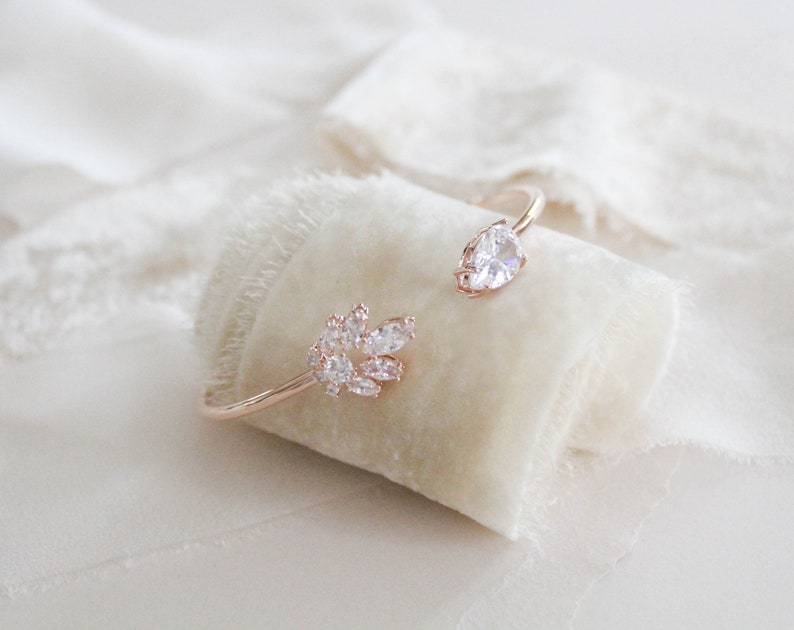 K&ouml;nnte beinhalten: Ein ros&eacute;goldenes Armband mit einem Cluster aus klaren Zirkonia-Steinen an einem Ende und einem einzelnen birnenf&ouml;rmigen Zirkonia-Stein am anderen Ende.