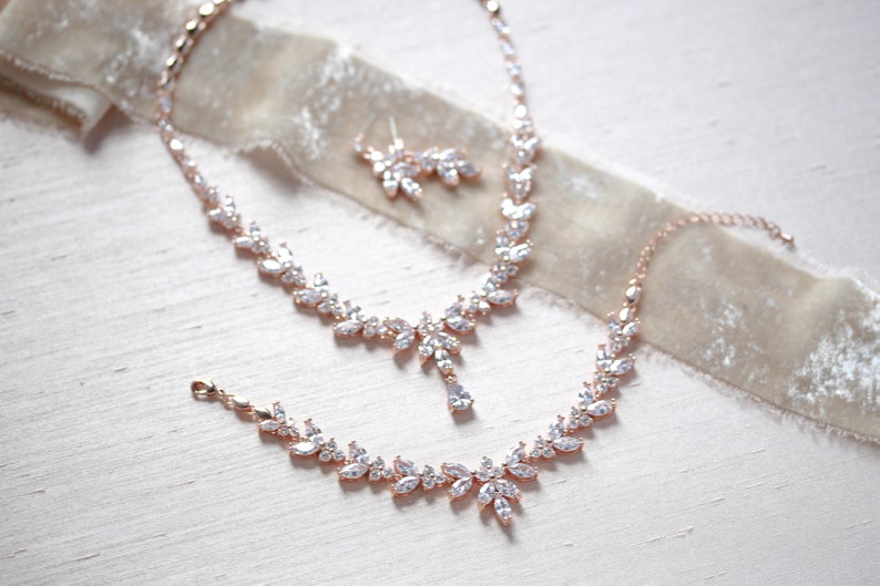 May include: A rose gold bridal jewellery set with a necklace, bracelet and earrings. The set features a delicate leaf design with clear cubic zirconia stones.