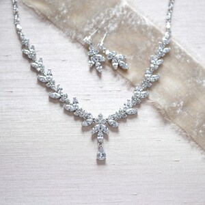 May include: A silver necklace and earrings set with a teardrop pendant and clear crystal accents. The jewellery is displayed on a light beige fabric background.