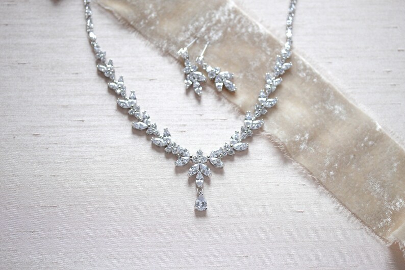 May include: A silver necklace and earrings set with a teardrop pendant and clear crystal accents. The jewellery is displayed on a light beige fabric background.