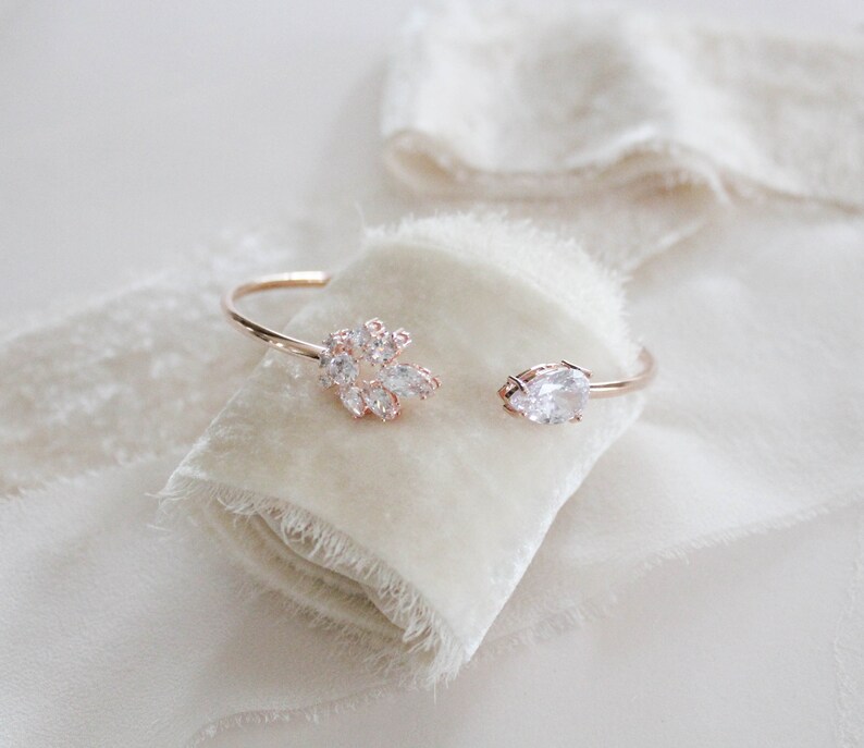 K&ouml;nnte beinhalten: Ein zartes Armband aus Ros&eacute;gold mit einem Cluster aus klaren Zirkonia-Steinen an einem Ende und einem einzelnen, gr&ouml;&szlig;eren birnenf&ouml;rmigen Stein am anderen.