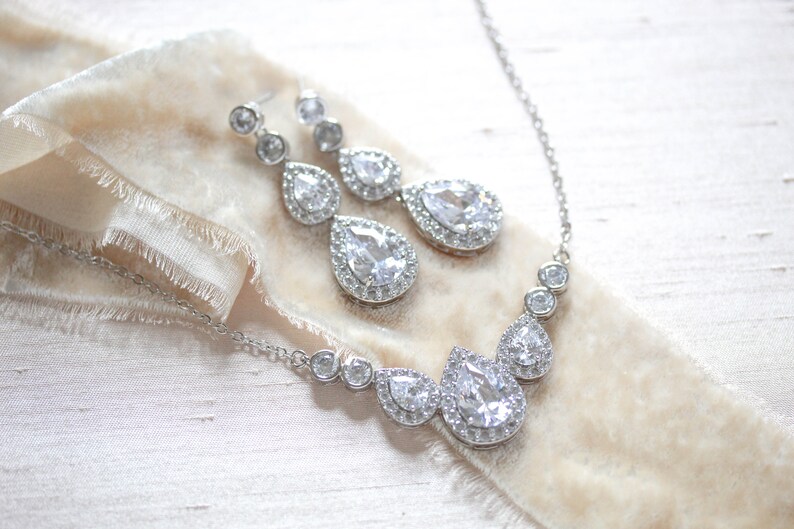 Pu&ograve; includere: Un set di collana e orecchini in argento con cristalli a forma di goccia d'acqua circondati da cristalli pi&ugrave; piccoli.