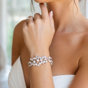 May include: A rose gold tone bracelet featuring a floral-inspired arrangement of clear marquise and round cut crystals interspersed with small white round beads. The bracelet has a flexible metal band design that wraps around the wrist.