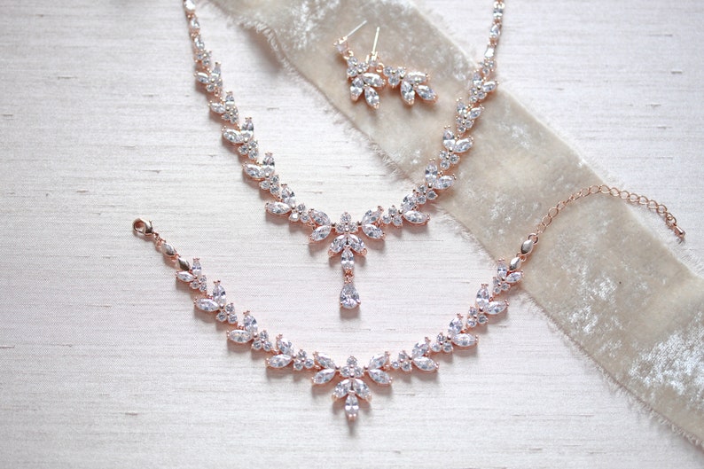 May include: A rose gold bridal jewellery set with a necklace, bracelet and earrings. The set features a delicate design with clear cubic zirconia stones.