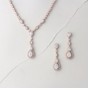 May include: A rose gold necklace and earrings set with pear-shaped clear crystals. The necklace has a long pendant with multiple crystals.