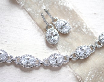May include: A silver bracelet and earrings set with oval-shaped clear stones surrounded by smaller stones. The jewelry is on a white background.