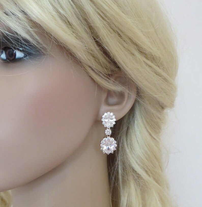 May include: Rose gold dangle earrings with clear cubic zirconia stones. The earrings feature a cluster of stones at the top and a larger oval-shaped stone at the bottom.