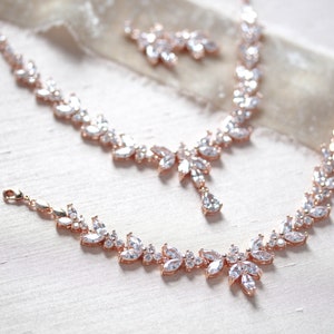May include: A rose gold bridal jewellery set with a necklace, bracelet and earrings. The jewellery features a delicate leaf design with clear cubic zirconia stones.