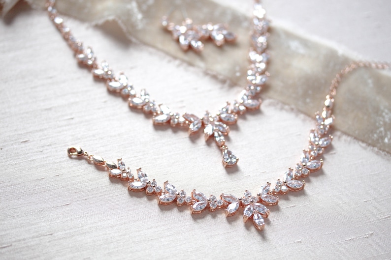 May include: A rose gold bridal jewellery set with a necklace, bracelet and earrings. The jewellery features a delicate leaf design with clear cubic zirconia stones.