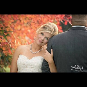 May include: A bride in a white strapless wedding dress with a sparkling necklace leans her head on the shoulder of the groom, who is wearing a gray suit. The background is a blurred image of fall foliage.
