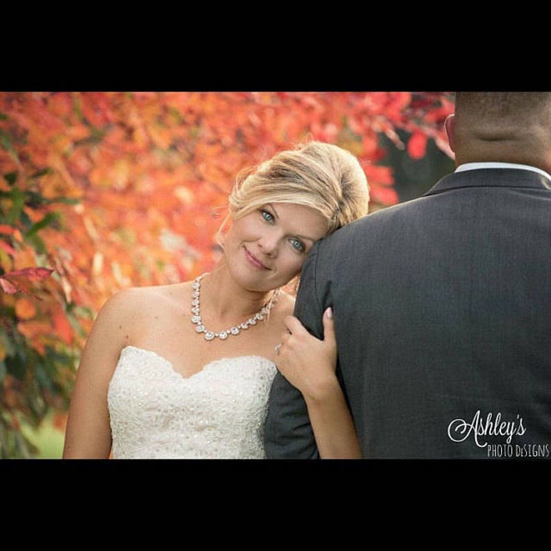 May include: A bride in a white strapless wedding dress with a sparkling necklace leans her head on the shoulder of the groom, who is wearing a gray suit. The background is a blurred image of fall foliage.