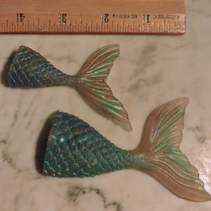 May include: Two mermaid tail ornaments with a green and blue iridescent finish. The tails are made of a translucent material with glitter embedded in the scales.