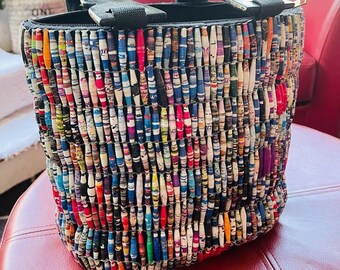 May include: A colorful tote bag made from rolled up pieces of paper. The bag has a black handle and a zipper closure.