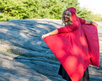 May include: A woman wearing a red hooded cape with a long, flowing design. The cape is made of a soft, lightweight fabric and has a relaxed fit. The woman is standing on a rocky surface with a green background.
