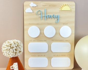 May include: A wooden birthday board with the name "Henry" in blue script, featuring spaces for "Today I Am," "Date," and "Height." Includes a yellow sun, white clouds, and a brown party hat with a "1."